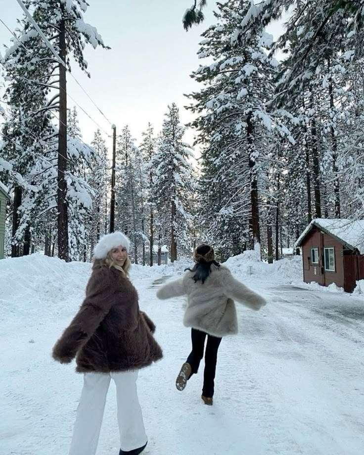 Two people in winter coats joyfully walk on a snowy road, surrounded by snow-laden trees and a cozy cabin. Winter wonderland scene.