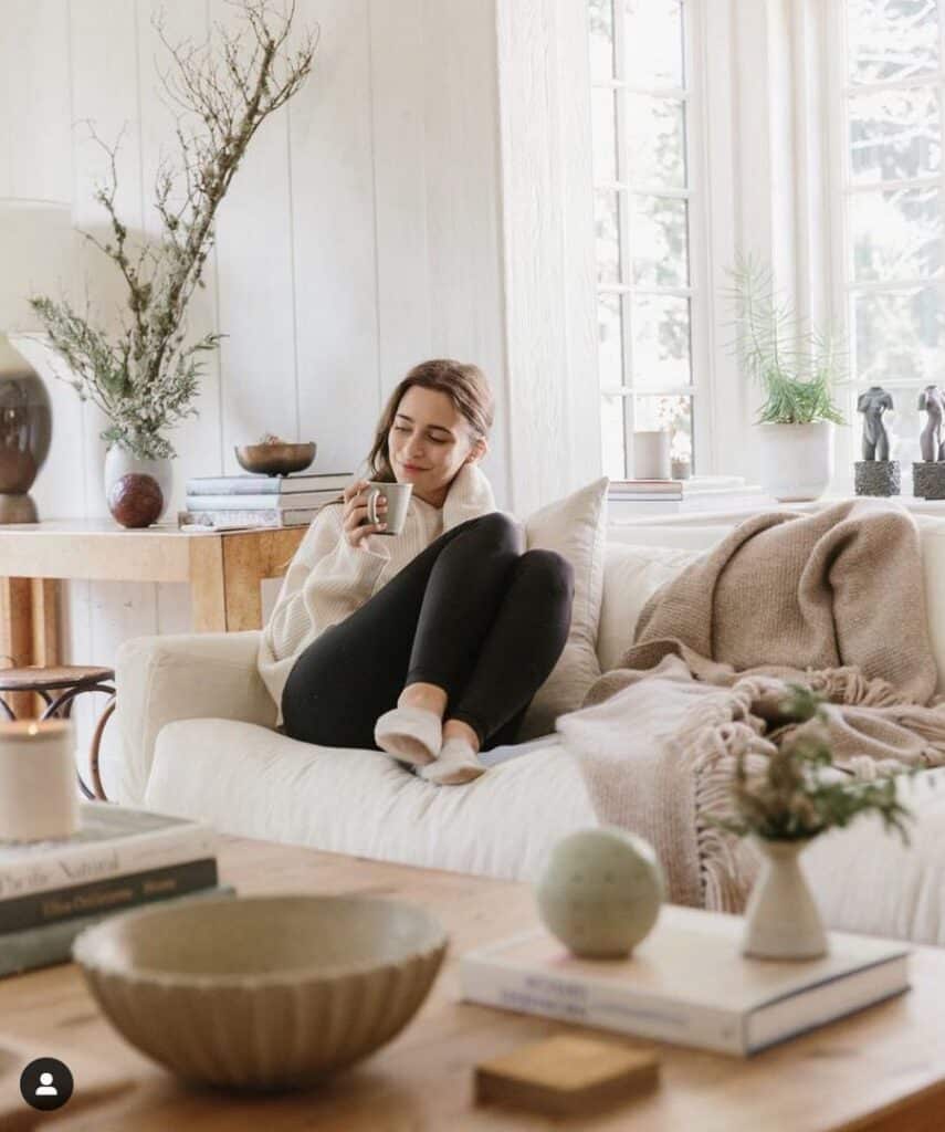 Cozy living room scene with a woman relaxing on a couch, sipping coffee, surrounded by books and plants by a bright window.