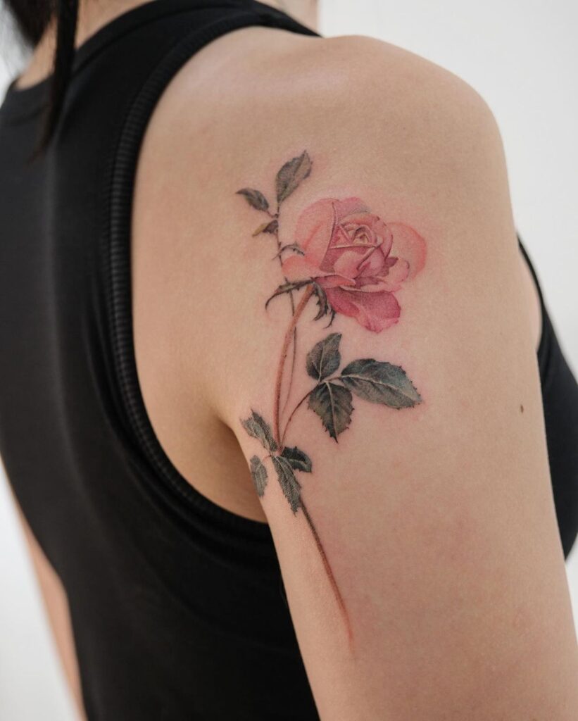 A detailed pink rose tattoo on a person's upper arm, blending artfully with their skin, wearing a black sleeveless top.