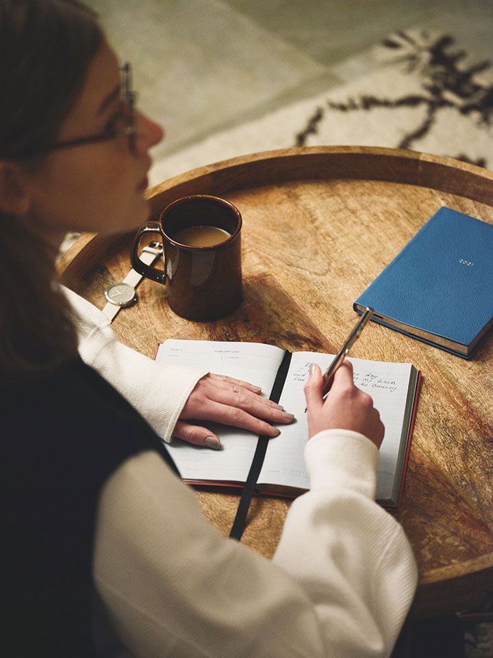 33184f5e319ed43107a8f598d95f36fe Person writing notes in a journal at a round wooden table with a coffee mug and a blue notebook nearby, creating a cozy workspace. | Sky Rye Design Person writing notes in a journal at a round wooden table with a coffee mug and a blue notebook nearby, creating a cozy workspace.