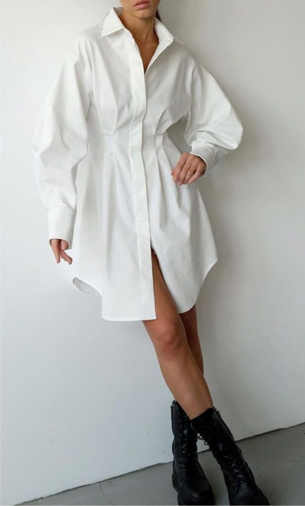 _ 34 Woman in a white shirt dress and black boots poses stylishly against a white wall, showcasing fashion-forward minimalist elegance. | Sky Rye Design Woman in a white shirt dress and black boots poses stylishly against a white wall, showcasing fashion-forward minimalist elegance.