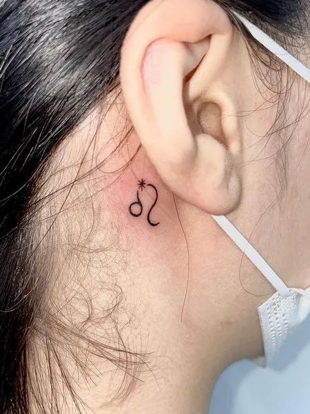 Small zodiac tattoo behind the ear featuring Leo symbol and a star, illustrating minimalist astrology-themed ink.