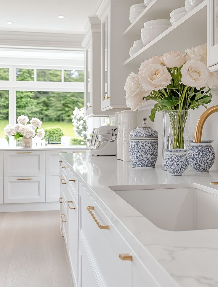 Elegant white kitchen with gold accents, marble countertops, and vases of fresh flowers, offering a bright and luxurious ambiance.