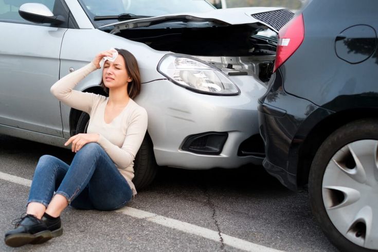 Woman sitting injured after car accident, holding head with cloth, two damaged cars in background.