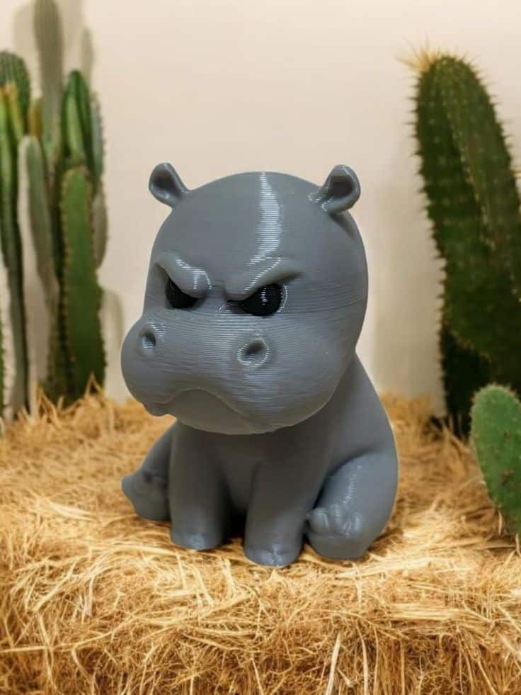 Cute grey 3D-printed hippo with an angry expression, sitting on hay with cactus plants in the background.