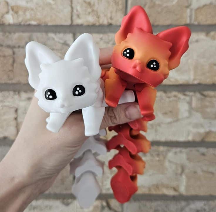 Hand holding two dragon-shaped fidget toys, one white and one red, against a brick wall background.