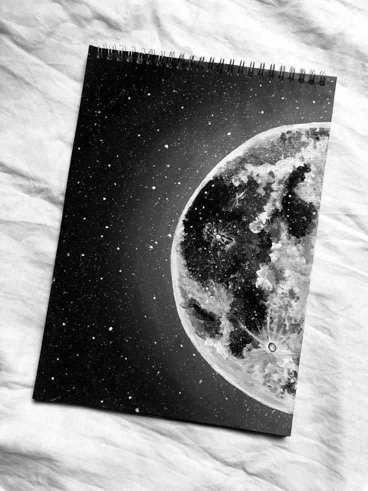 Black and white sketchbook drawing of the moon and stars on a spiral-bound notebook, placed on a textured white surface.