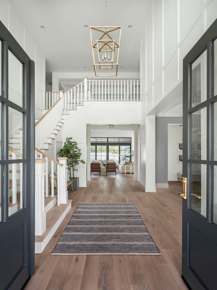 Spacious modern entryway with wooden floors, elegant staircase, and stylish lighting, leading to a bright living area through glass doors.