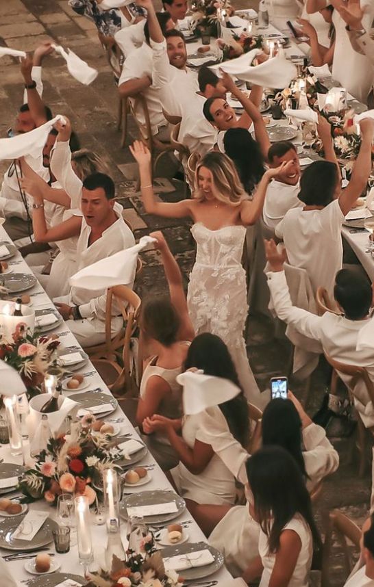 _ 4 Elegant wedding reception with guests waving napkins, celebrating around a beautifully decorated table. Bride in lace gown joins in joyfully. | Sky Rye Design Elegant wedding reception with guests waving napkins, celebrating around a beautifully decorated table. Bride in lace gown joins in joyfully.