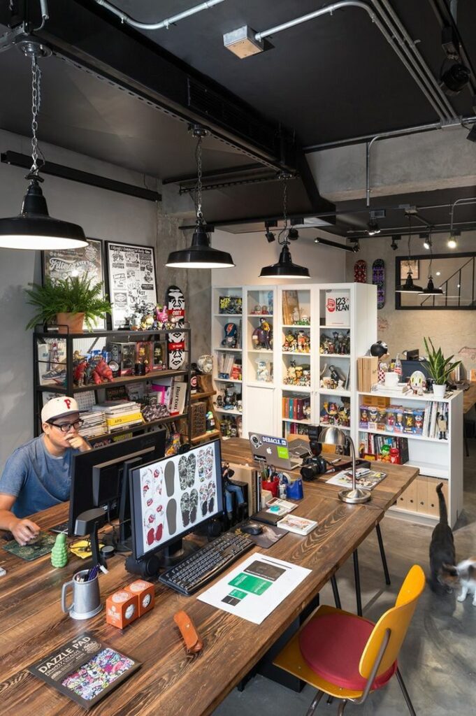 Creative office space with art, plants, and collectibles. Person working at a desk with computers, surrounded by vibrant decor.