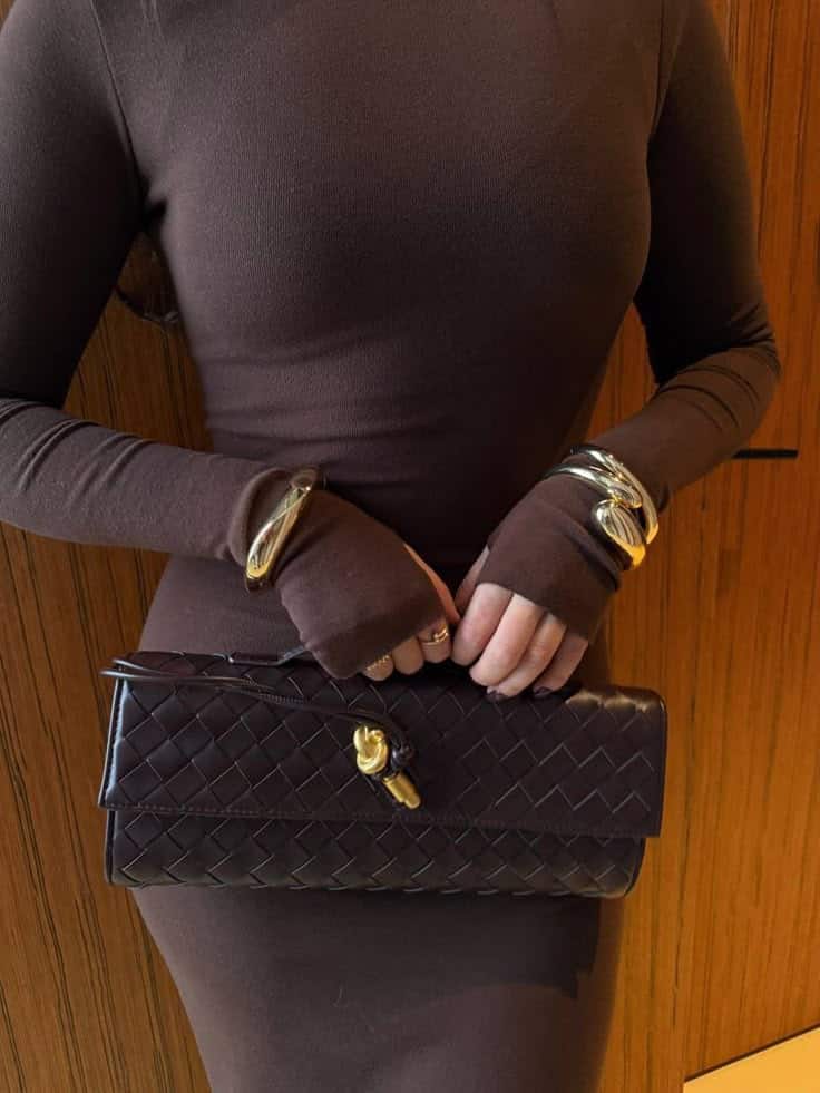 _ 40 Woman in brown dress holding a woven leather clutch, accessorized with gold bangles, standing against a wooden background. | Sky Rye Design Woman in brown dress holding a woven leather clutch, accessorized with gold bangles, standing against a wooden background.