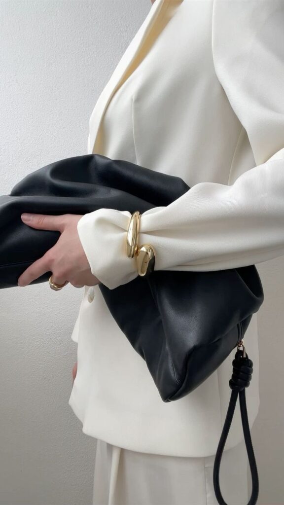 _ 41 Person in white suit holding a large black leather clutch, accented with gold bracelets, showcasing elegant fashion style. | Sky Rye Design Person in white suit holding a large black leather clutch, accented with gold bracelets, showcasing elegant fashion style.