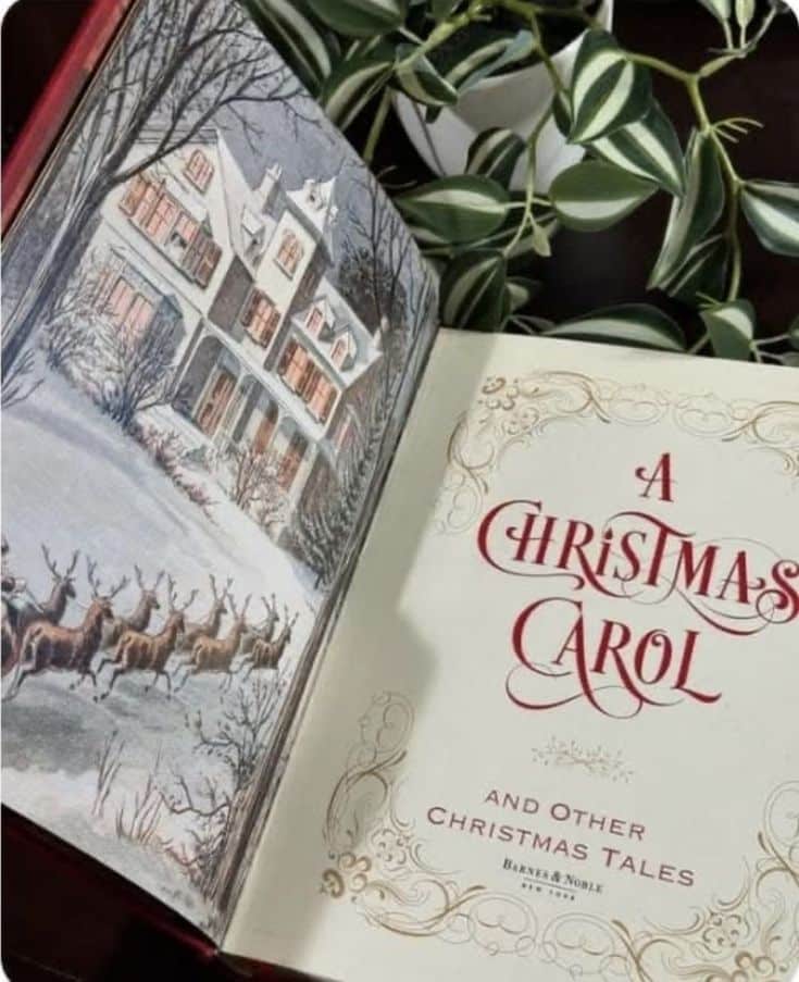 Open A Christmas Carol book with snowy illustration and reindeer, surrounded by green leaves on a wooden table.