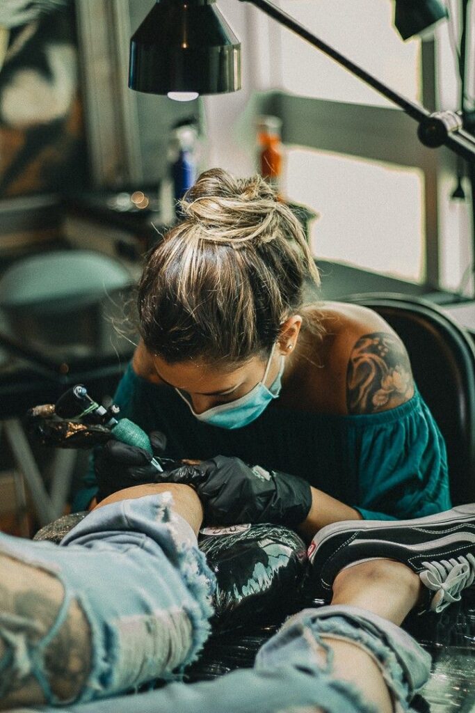 42147c046ea32a8f295887a9daf729e4 Tattoo artist in a mask and gloves working on a client's arm in a studio, showcasing detailed ink art and professional care. | Sky Rye Design Tattoo artist in a mask and gloves working on a client's arm in a studio, showcasing detailed ink art and professional care.