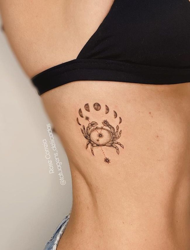 Side torso tattoo of a crab with moon phases, stars, and delicate line work, symbolizing astrology and cosmic themes, in black ink.
