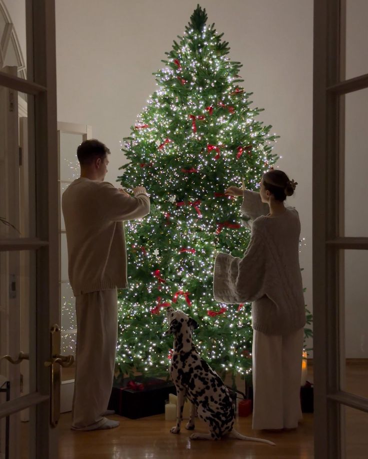 Cozy Christmas scene with couple decorating a lit tree, red bows glimmering, and Dalmatian watching in festive home setting.