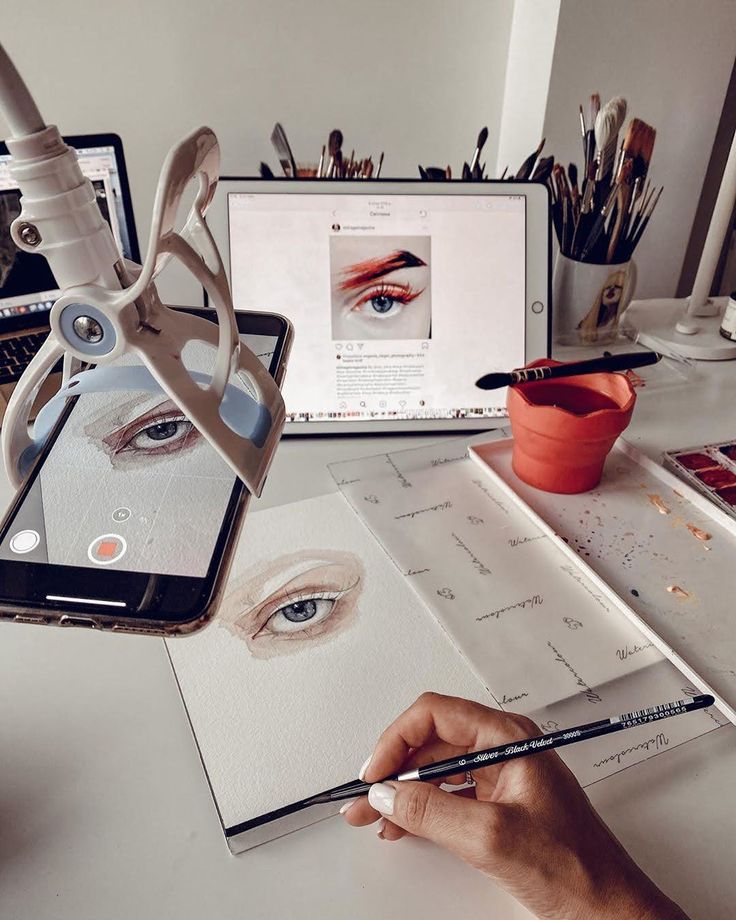 Artist sketching an eye with a pencil, captured on smartphone, tablet in background, brushes and paints scattered on desk.