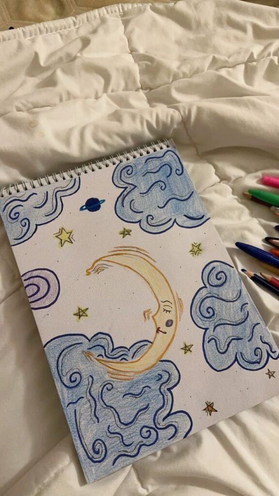 Illustration on sketchbook of crescent moon, stars, and clouds with colored pens on a cozy white blanket.