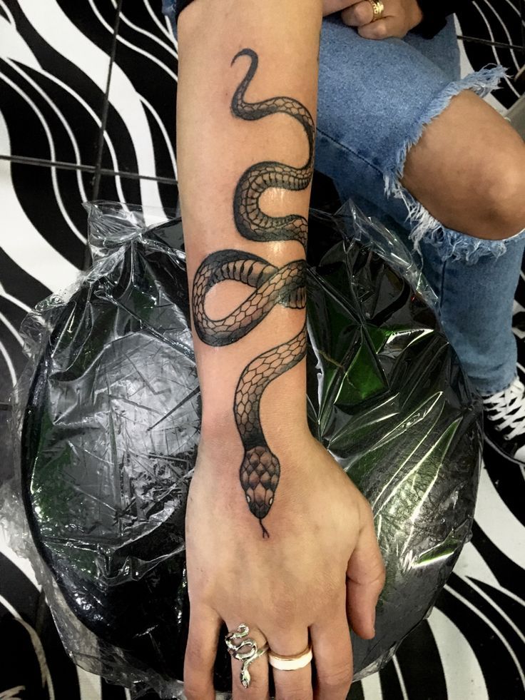 Snake tattoo design winding around a forearm, showcasing intricate scales and details. Denim jeans and rings are visible in the background.