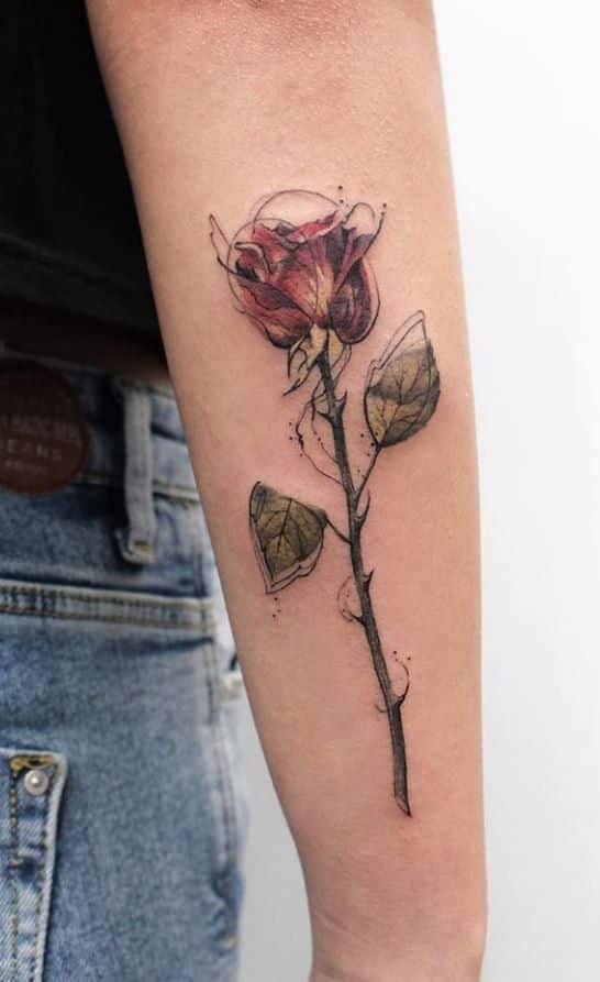 Watercolor rose tattoo on forearm, featuring realistic flower and leaves, in jeans background.