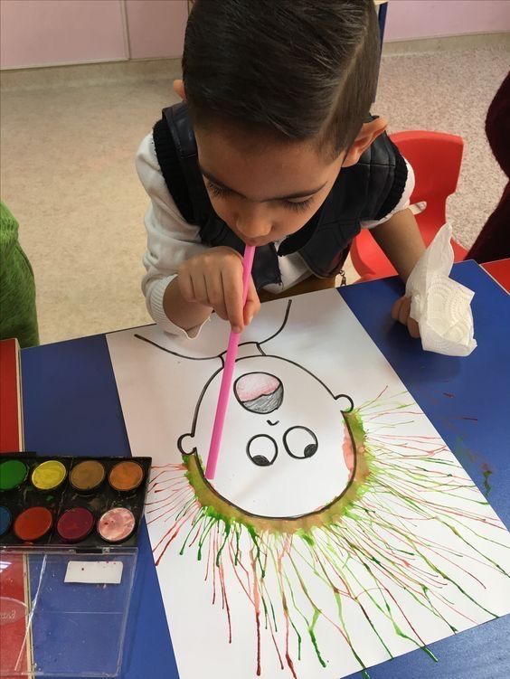 Child creating art by blowing paint through a straw onto a paper with a face outline, using vibrant colors on a classroom table.