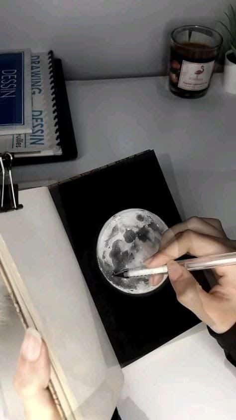 Artist sketching a realistic moon in a notebook on a desk, with drawing books and a candle in the background.