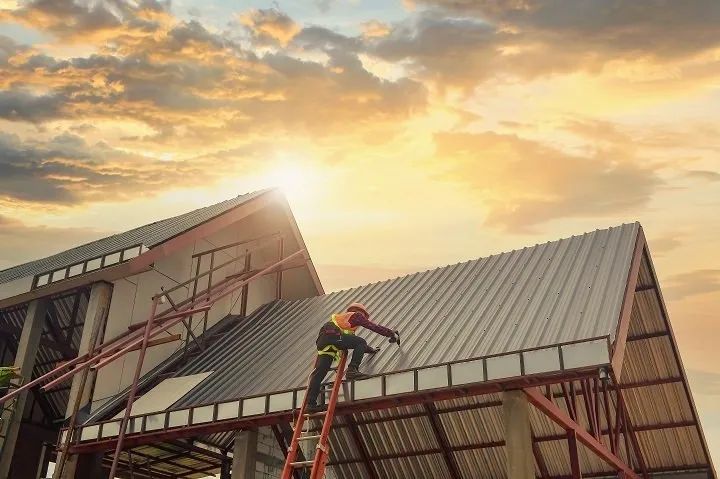 _ 5 Worker installing metal roof panels on a house under construction at sunset. | Sky Rye Design Worker installing metal roof panels on a house under construction at sunset.