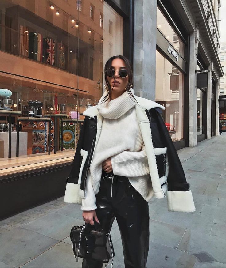 Woman in chic winter fashion with sunglasses, white turtleneck, leather jacket and pants, standing on urban sidewalk.