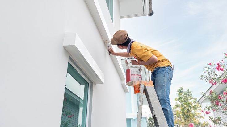 HomeOrHousePaintingWorkConsistOfPainterManOr Person painting house exterior on ladder with paintbrush and bucket, wearing casual attire under clear sky. Home improvement project. | Sky Rye Design Person painting house exterior on ladder with paintbrush and bucket, wearing casual attire under clear sky. Home improvement project.