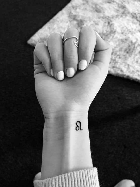 Minimalist wrist tattoo of the Leo zodiac symbol, shown with a stylish finger ring on a monochrome background.