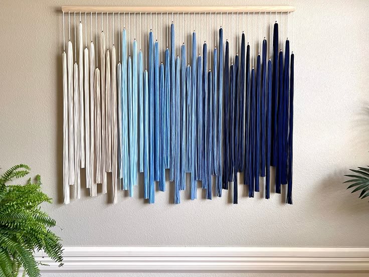 Colorful yarn wall hanging with shades of blue, cream, and white on a beige wall. Green plants frame the artistic display.