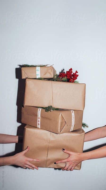 Hands holding a stack of brown paper-wrapped gifts adorned with festive greenery and red berries against a plain white background.