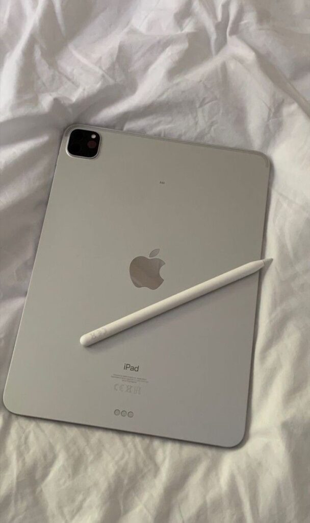 Silver iPad with an Apple Pencil on top, laid on white bedding, showcasing modern technology and sleek design.