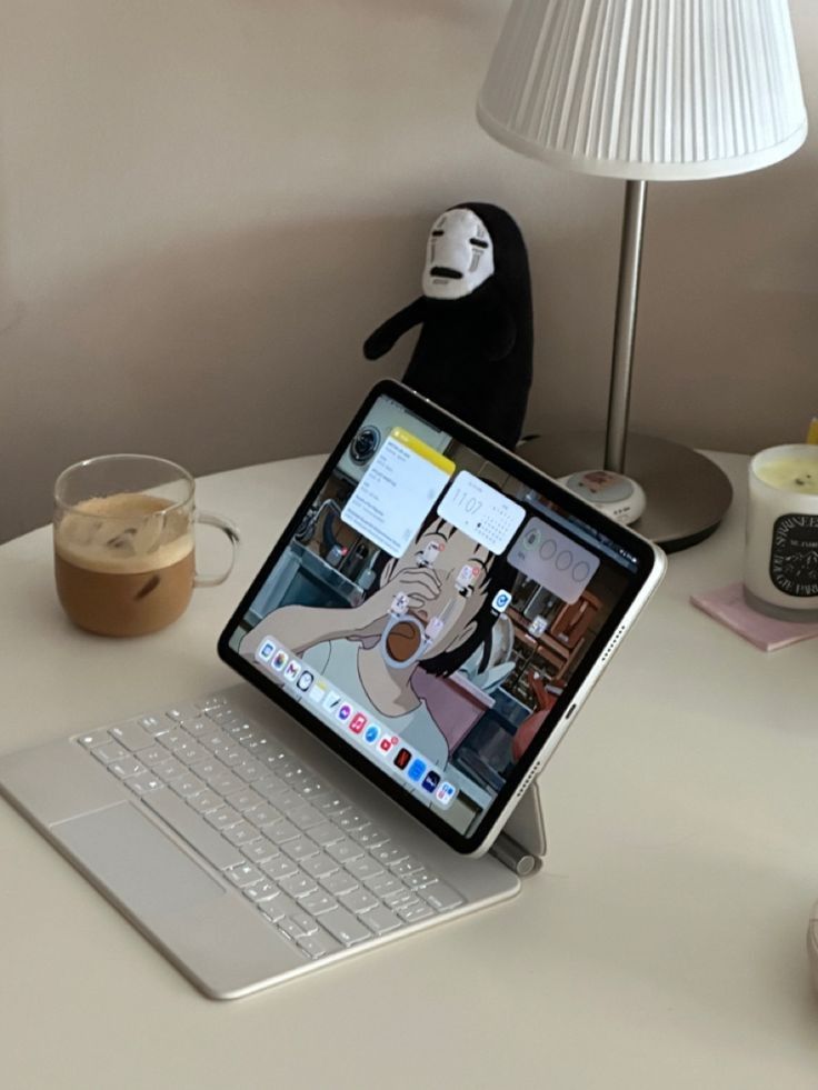 Tablet on a desk with a keyboard, featuring an anime wallpaper, next to a coffee, a lamp, and a plush toy for a cozy workspace vibe.