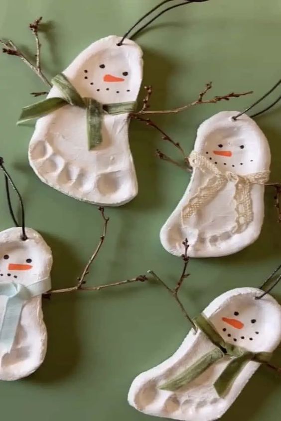 Clay snowman ornaments made from footprints, decorated with ribbons and twigs for arms, on a green background. Perfect DIY holiday decor.