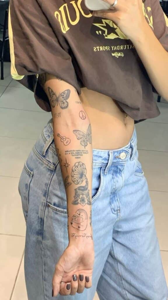 559a128f-b901-48b9-8387-f61392cff2d0 Person showing arm tattoos, including butterflies and symbols, wearing a brown cropped top and light jeans. | Sky Rye Design Person showing arm tattoos, including butterflies and symbols, wearing a brown cropped top and light jeans.