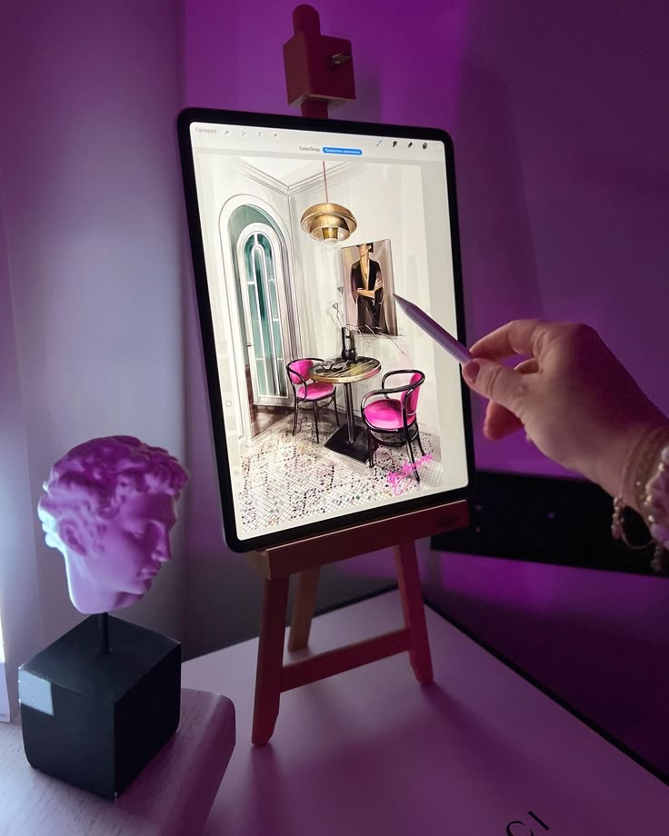 Hand using stylus on tablet displaying elegant room design with pink chairs, next to a sculpture bust under purple lighting.