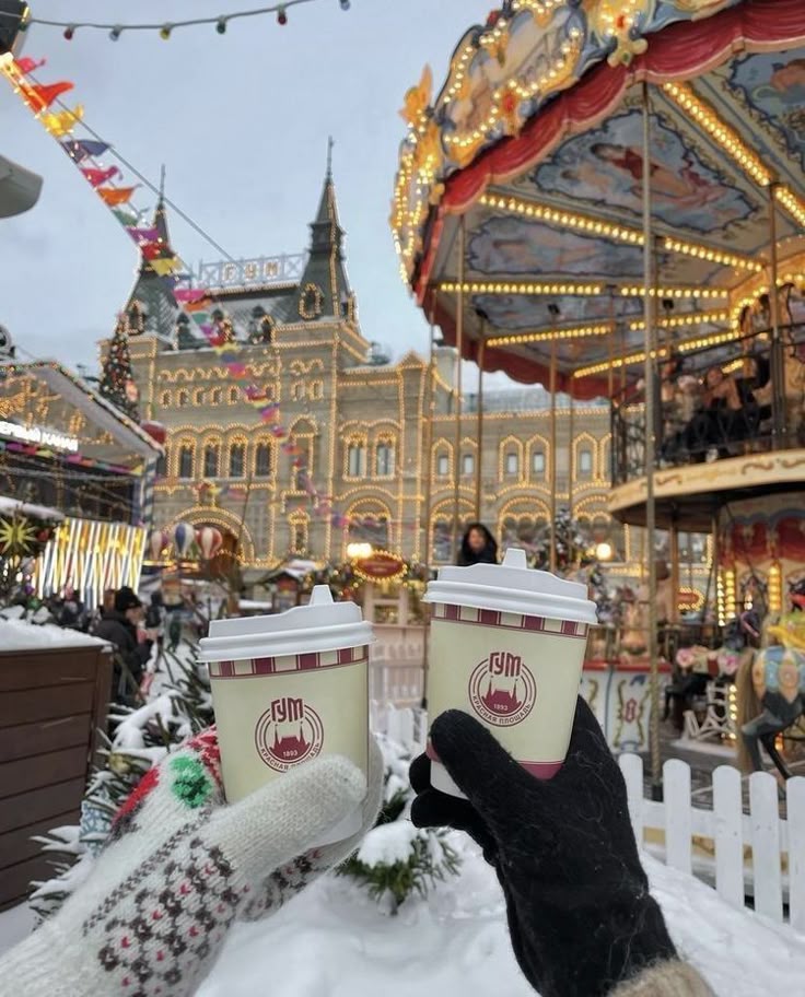 Gloved hands holding coffee cups in front of a snowy amusement park with a carousel and festive lights.
