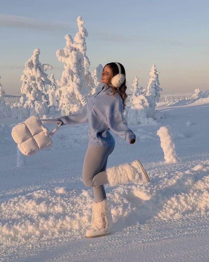 Woman in winter attire enjoys snowy landscape, wearing earmuffs and boots, holding a puffy bag, surrounded by snow-covered trees.
