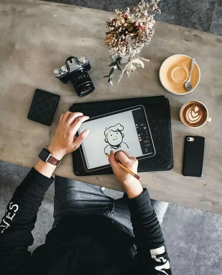 Artist sketching a cartoon on a tablet, with a camera, coffee, and flowers on a table. Creative workspace setup with digital art.