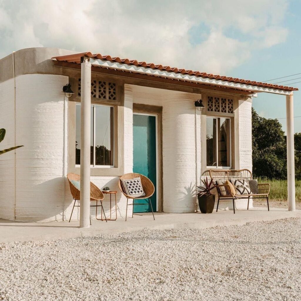 Small modern house with chairs on porch, gravel yard, blue door, sunny day, showcasing minimalist design and outdoor seating.