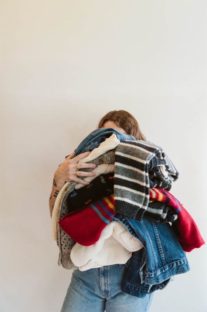 Person holding a large stack of colorful clothes, including sweaters and jeans, against a plain white background.