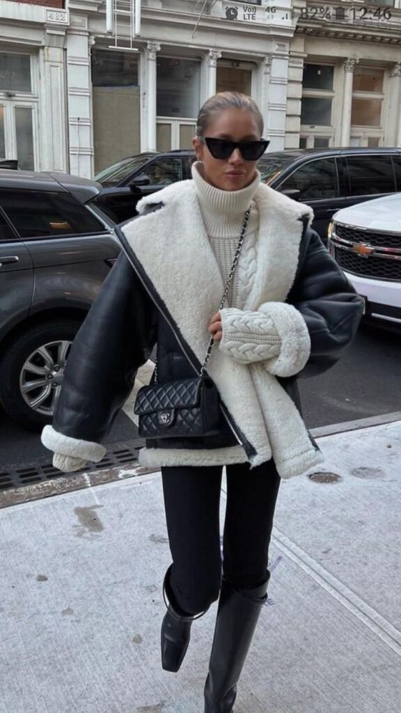 Stylish woman in winter fashion: shearling coat, sunglasses, black boots, and quilted handbag walking in city street.