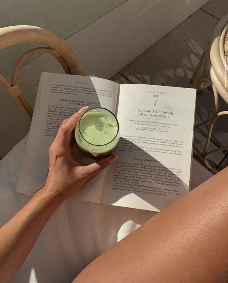 _ 6 Person reading a book on goal-setting holding a green smoothie, relaxing in the sunlight. | Sky Rye Design Person reading a book on goal-setting holding a green smoothie, relaxing in the sunlight.