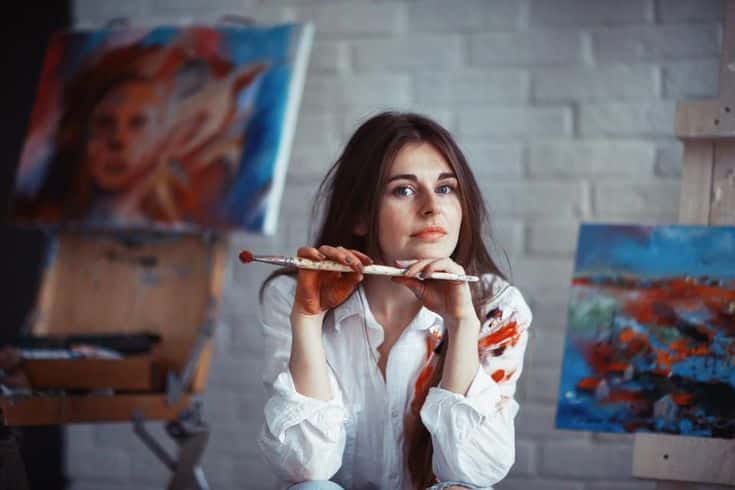 Artist in a studio surrounded by colorful paintings, holding paintbrushes and wearing a paint-splattered shirt, against a brick wall backdrop.