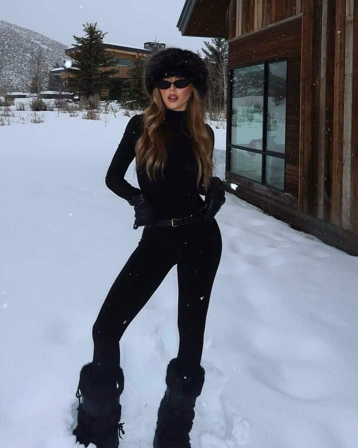 Stylish woman in black winter outfit poses outdoors in snowy landscape near wooden cabin.