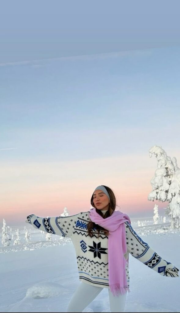 Woman in cozy sweater and pink scarf enjoying snowy winter landscape at sunset.