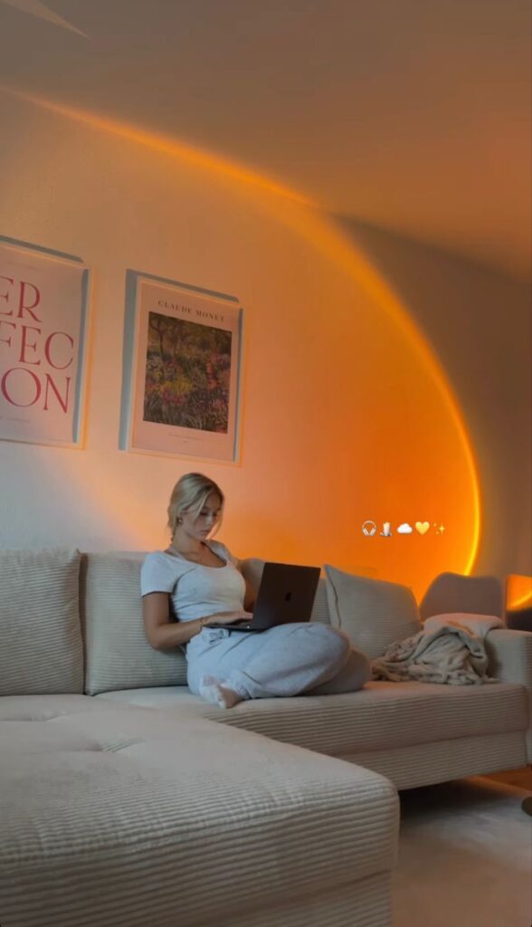Person relaxing on a sofa with a laptop, surrounded by warm lighting and modern wall art in a cozy living room.