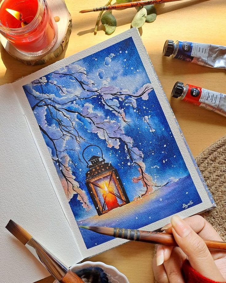 Watercolor painting of a lantern amidst snowy branches, surrounded by art supplies. Cozy, winter-themed artistic scene with vibrant colors.