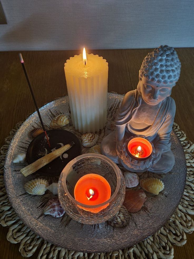 Tranquil setup with Buddha statue, lit candles, incense, and seashells on a decorative tray, creating a serene meditation corner.